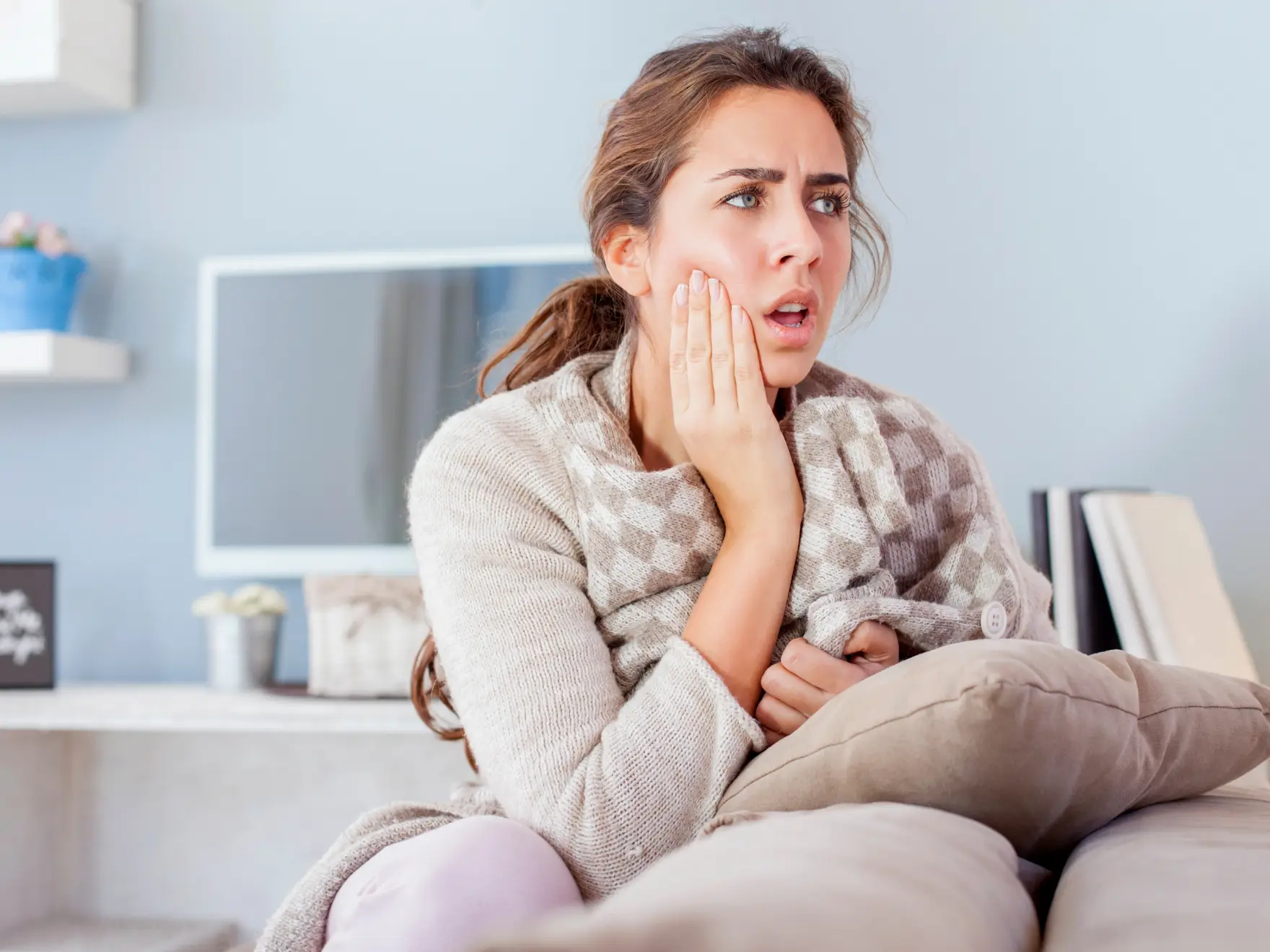 A woman sits on a couch, holding her cheek and looking worried.