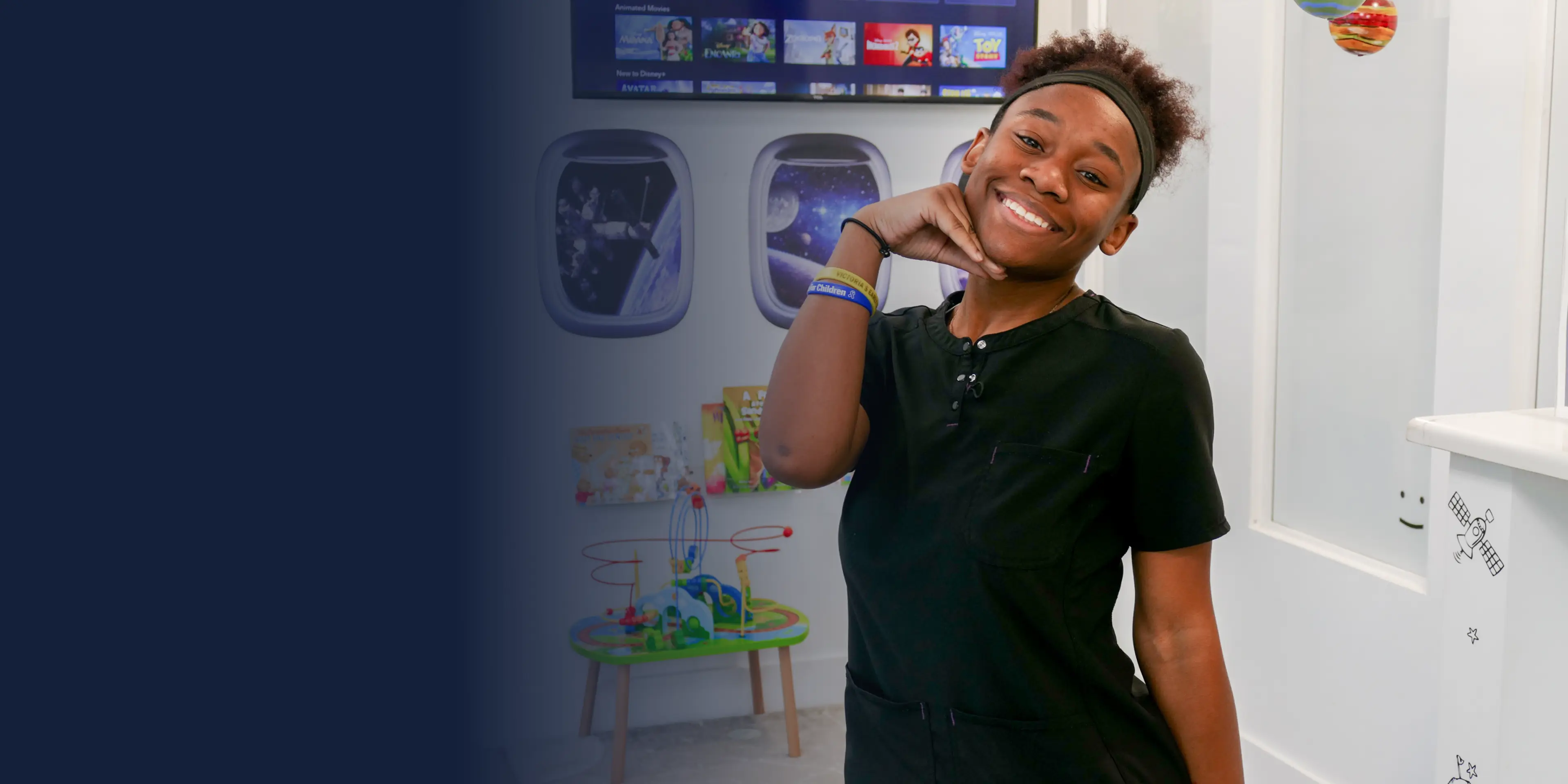 A person in black clothing stands indoors, smiling, with gaming or entertainment displays in the background.