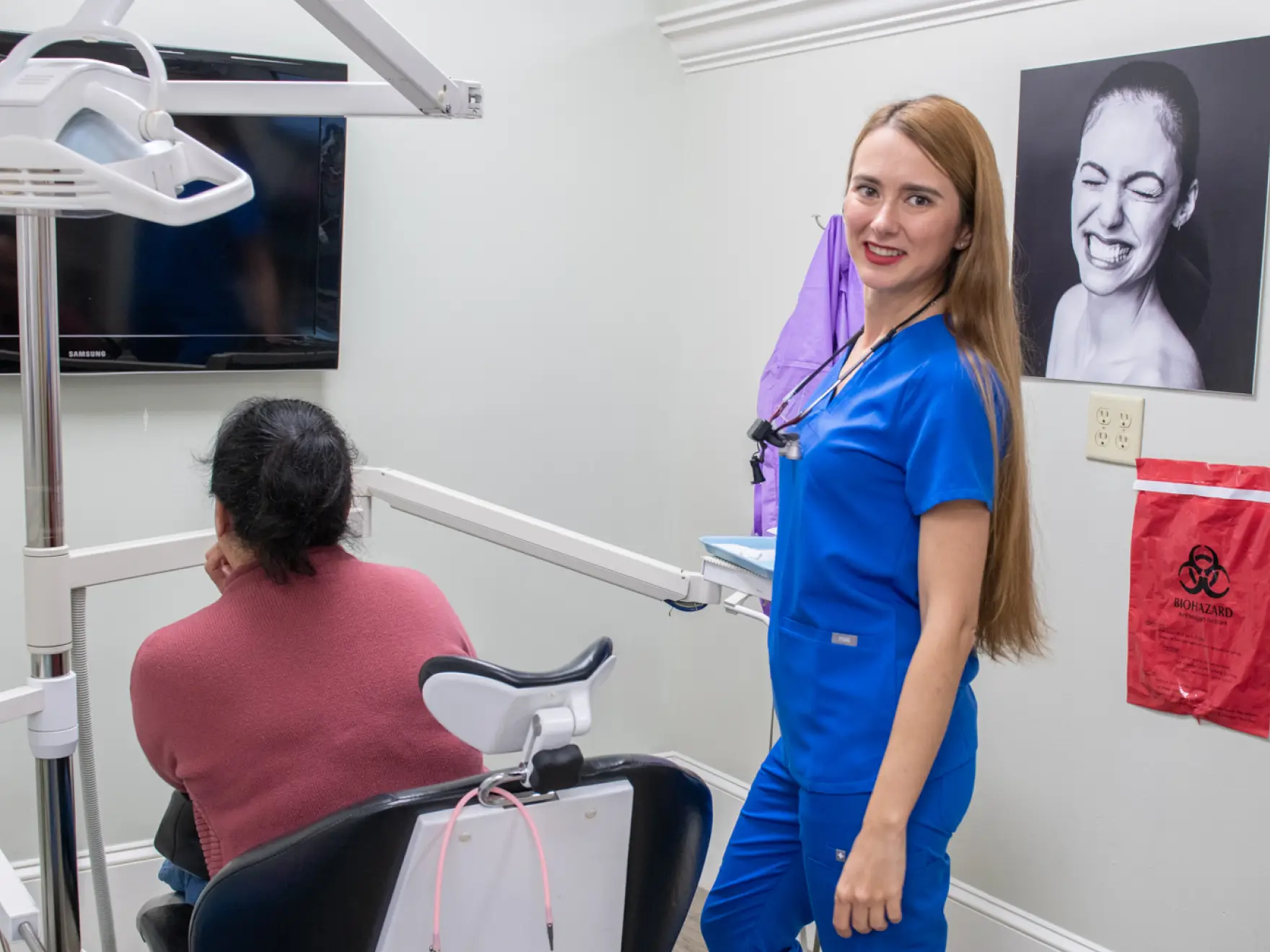 A dentist in blue scrubs smiles at the camera while a patient sits in the exam chair.