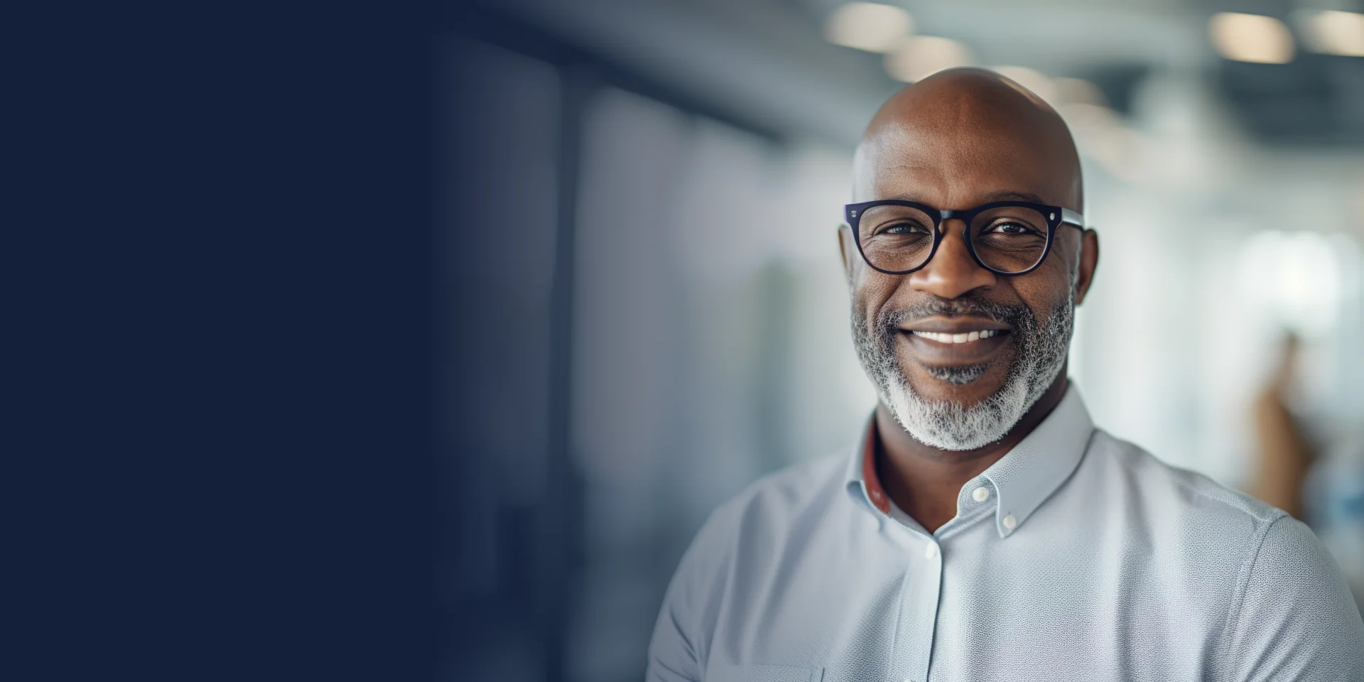 A man with glasses and a beard smiles, standing indoors.