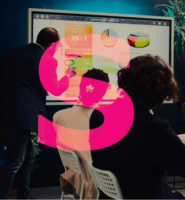 Business meeting with a presenter pointing at a large screen displaying business analytics graphs and charts while two attendees watch.