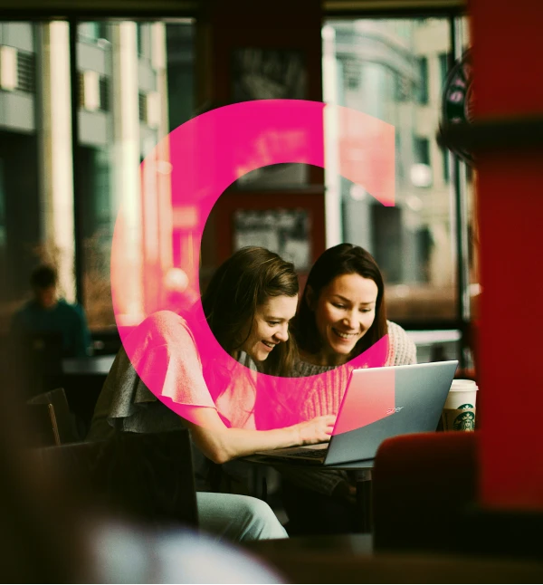 Two women smiling and looking at a laptop screen together in a cozy café setting.
