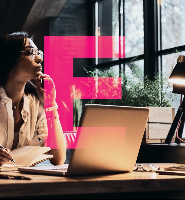 Woman with glasses sitting at a desk with a laptop and notebook, looking thoughtfully out a large window.