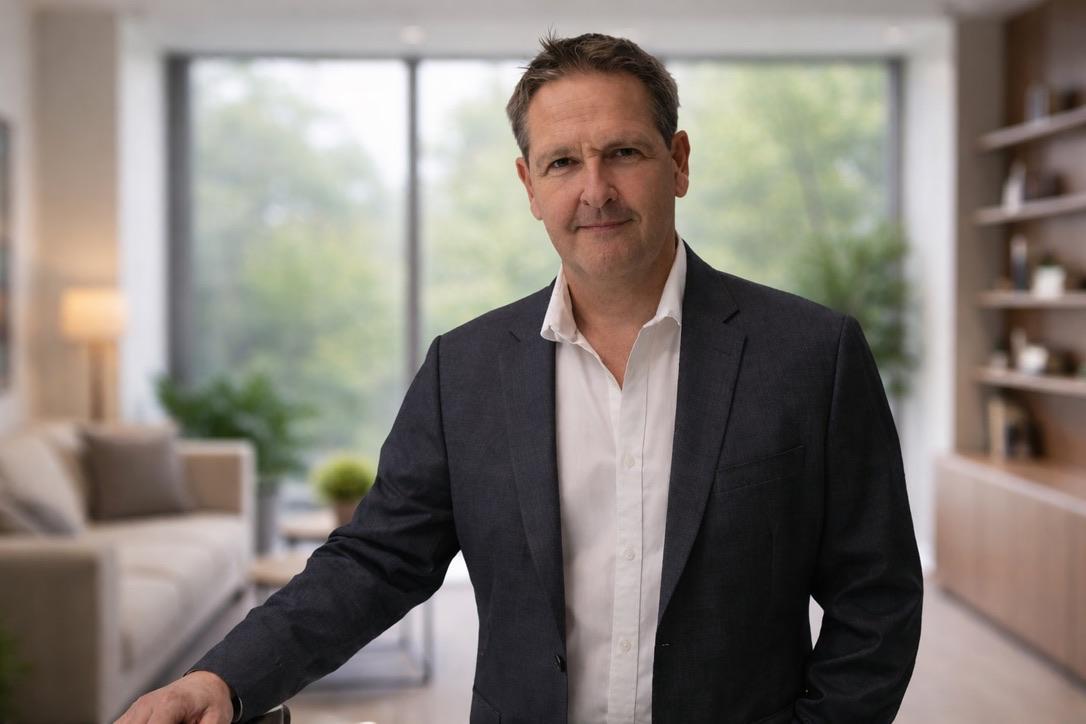 Young man in a blazer with arms crossed, standing indoors next to hanging industrial lamps and a staircase.
