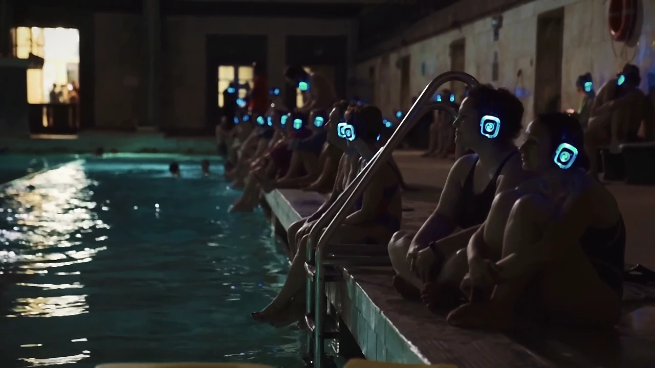 Fila de asistentes con auriculares iluminados sentados al borde de una piscina de noche