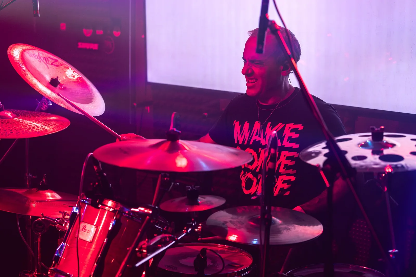 Batería sonriendo tras sus platillos y timbales, con camiseta serigrafiada, bajo luz rosa de escenario