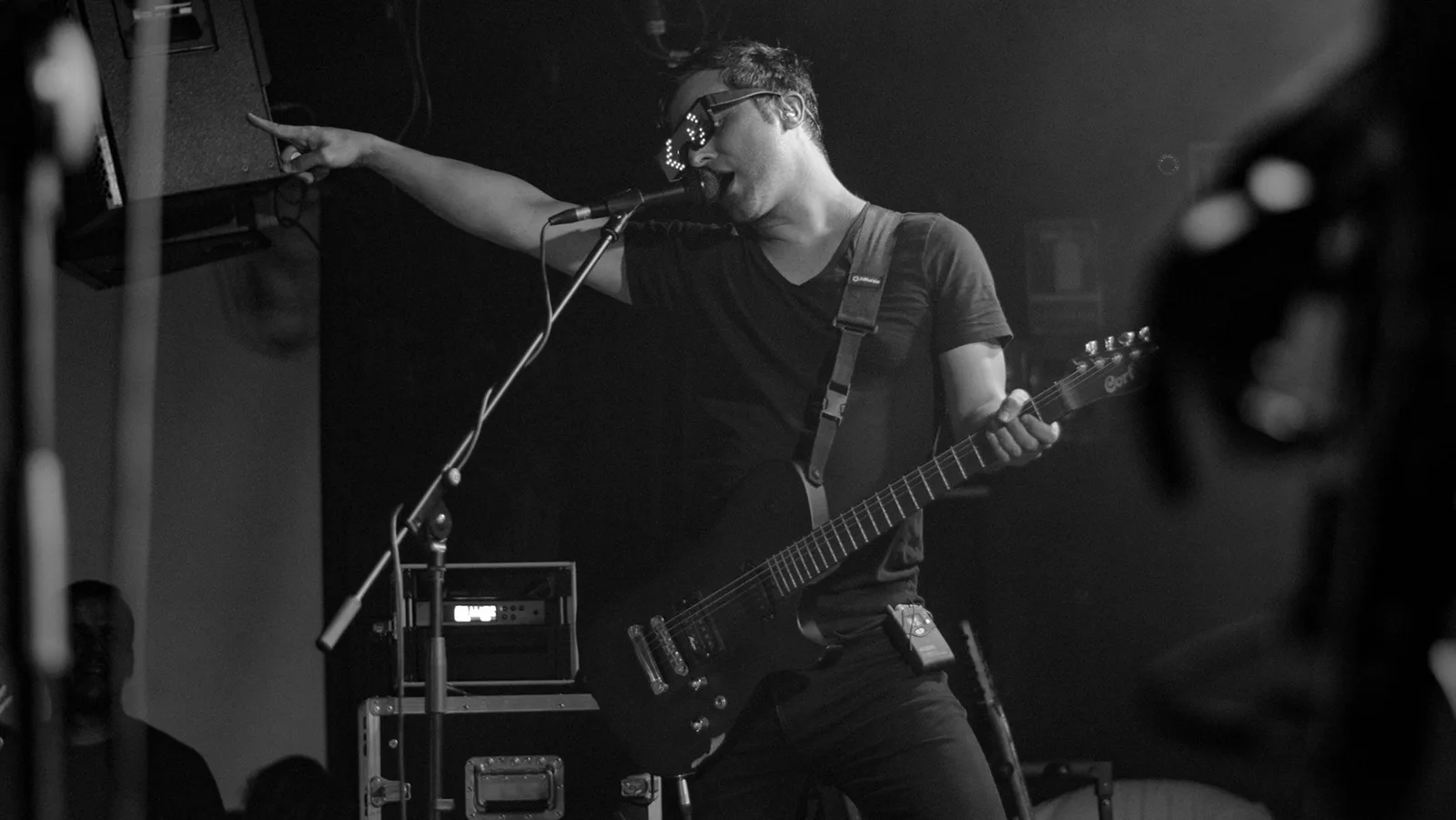 Imagen en blanco y negro de un cantante con gafas luminosas señalando mientras toca la guitarra