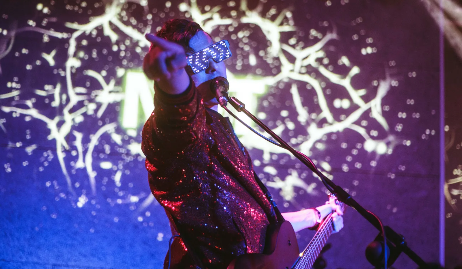 Cantante con gafas luminosas y chaqueta de lentejuelas señalando al público ante una proyección azulada
