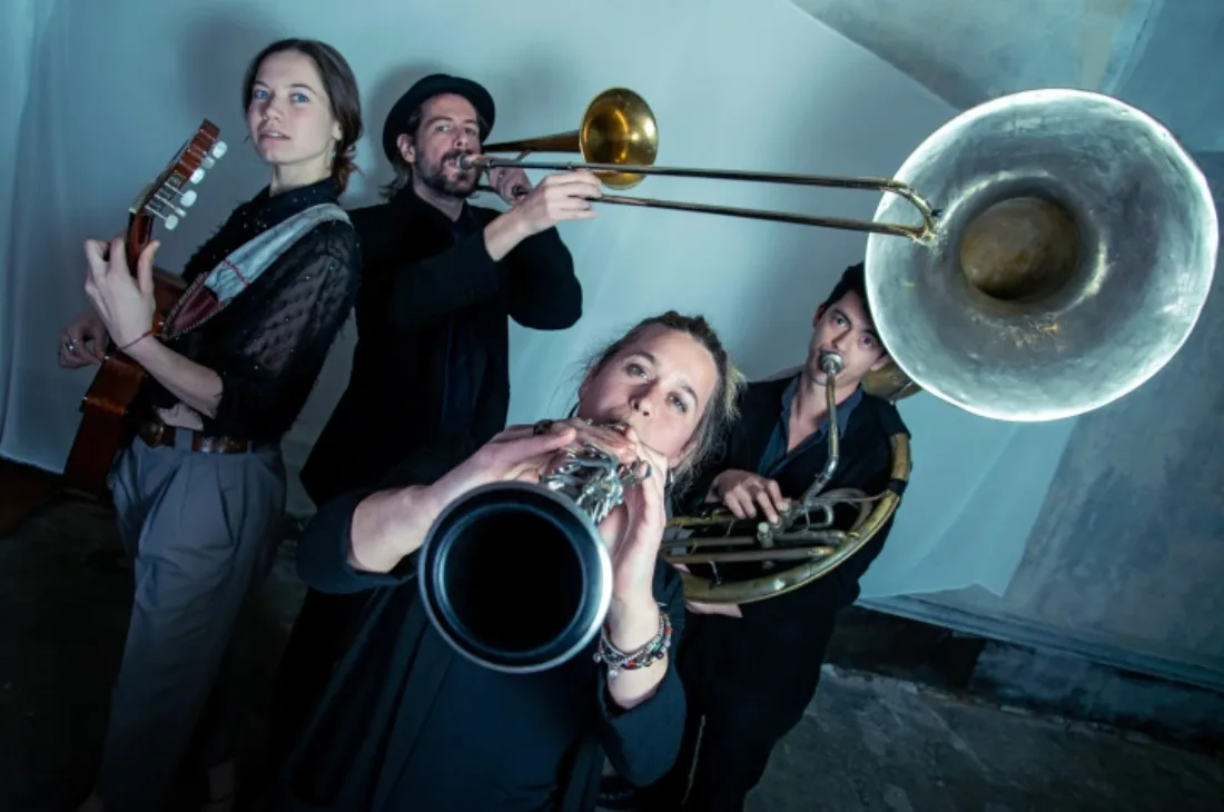 Cuatro músicos vistos en contrapicado con trombón, sousafón, un viento metálico y una guitarra al fondo
