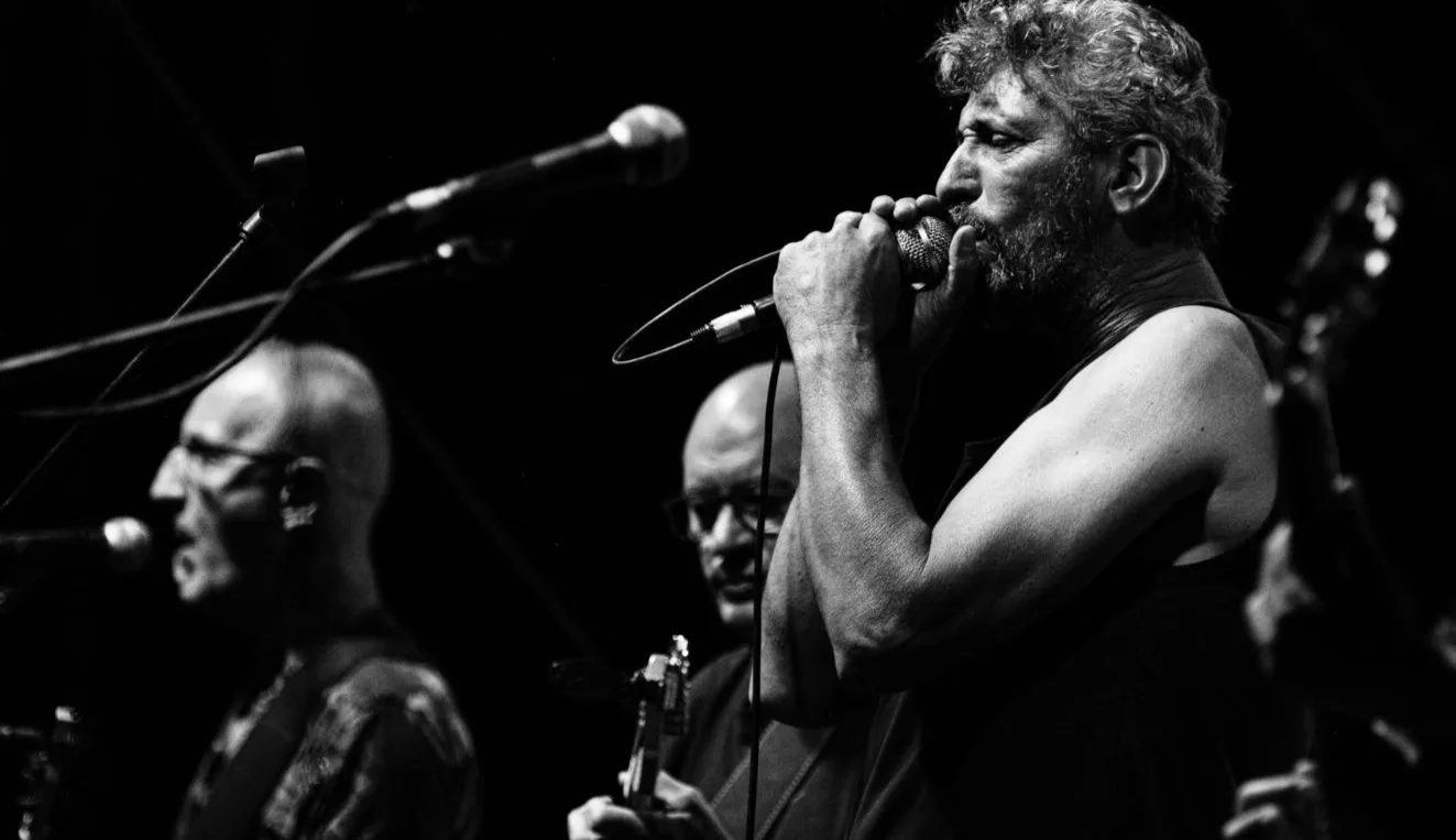 Cantante al micrófono en blanco y negro con dos músicos al fondo