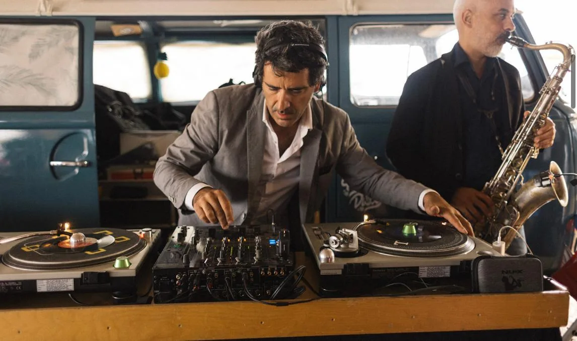 DJ mezclando con dos platos de vinilo y saxofonista tocando junto a una furgoneta clásica.