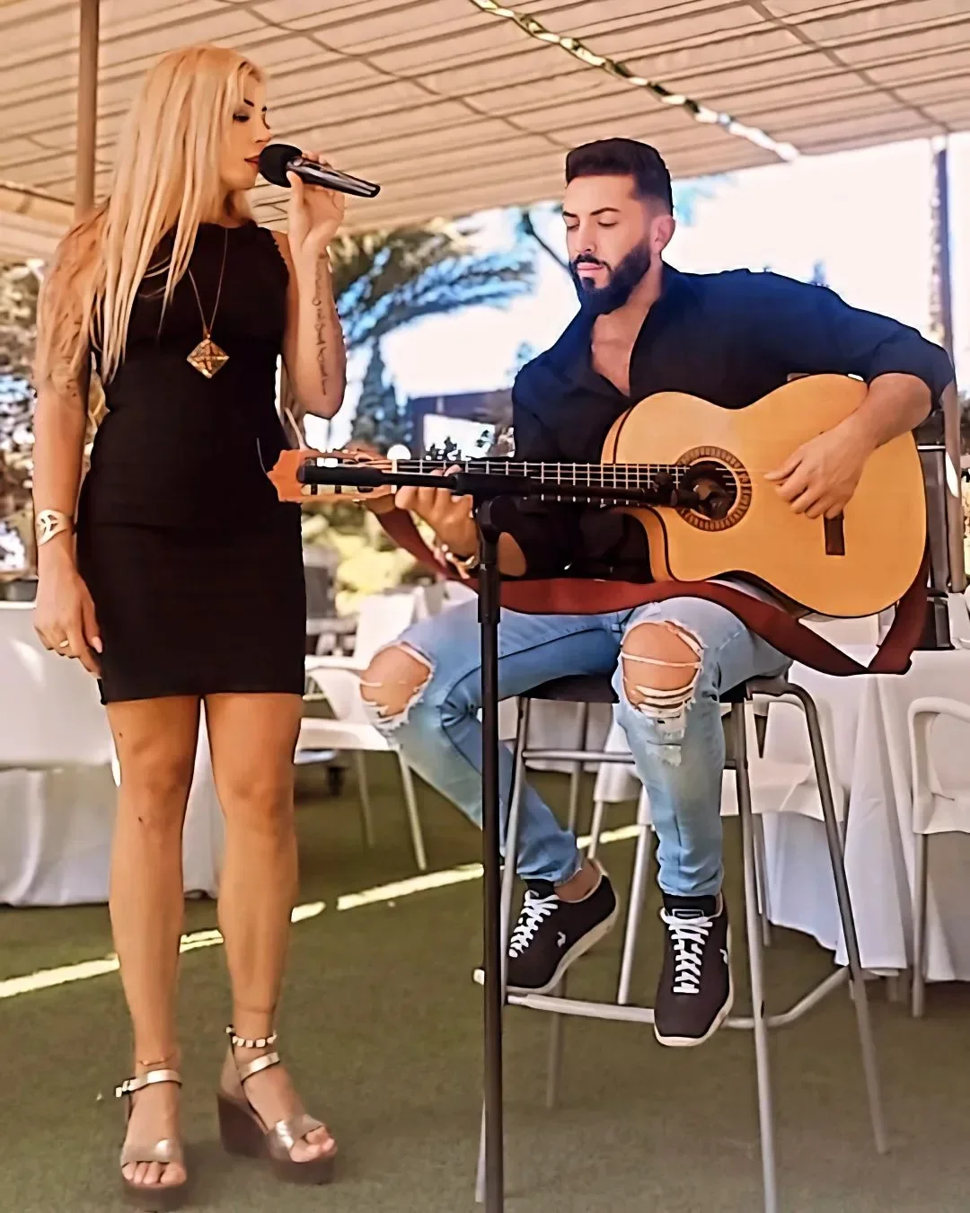 Dúo bajo una pérgola entre mesas vestidas de blanco: cantante de negro con micrófono y guitarrista sentado