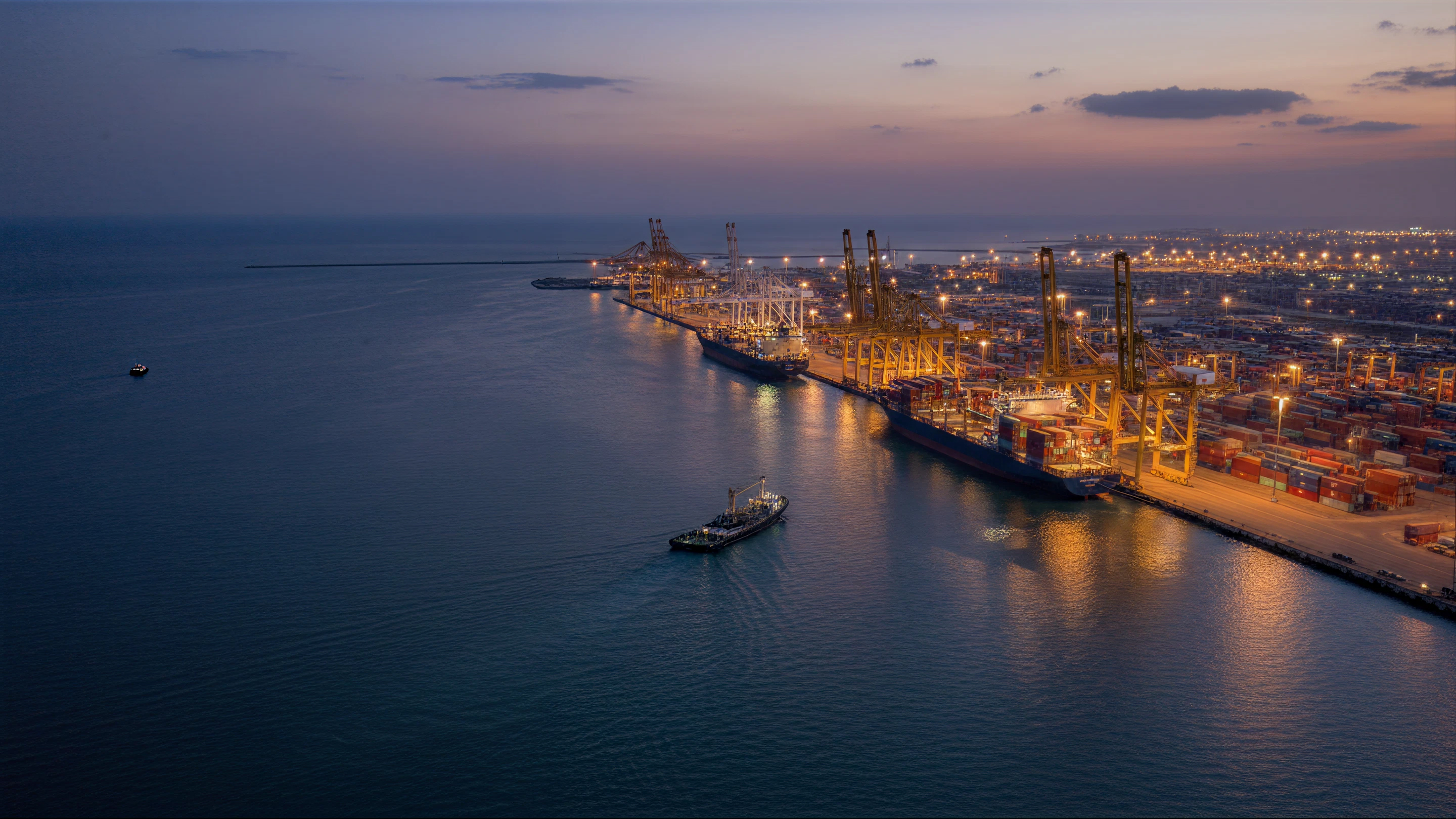 Cargo ship with containers guided by tugboats in a busy port with cranes and stacks of containers.