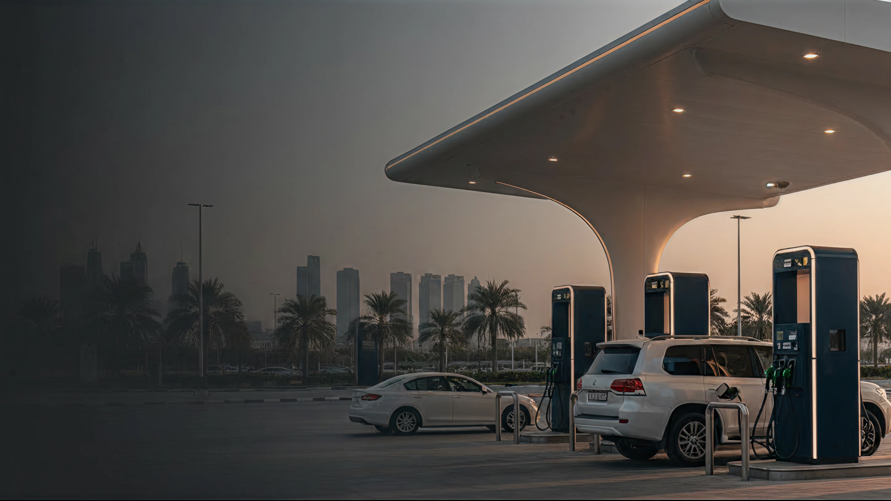 Two white cars refueling at a modern gas station with a city skyline and palm trees behind at dusk.