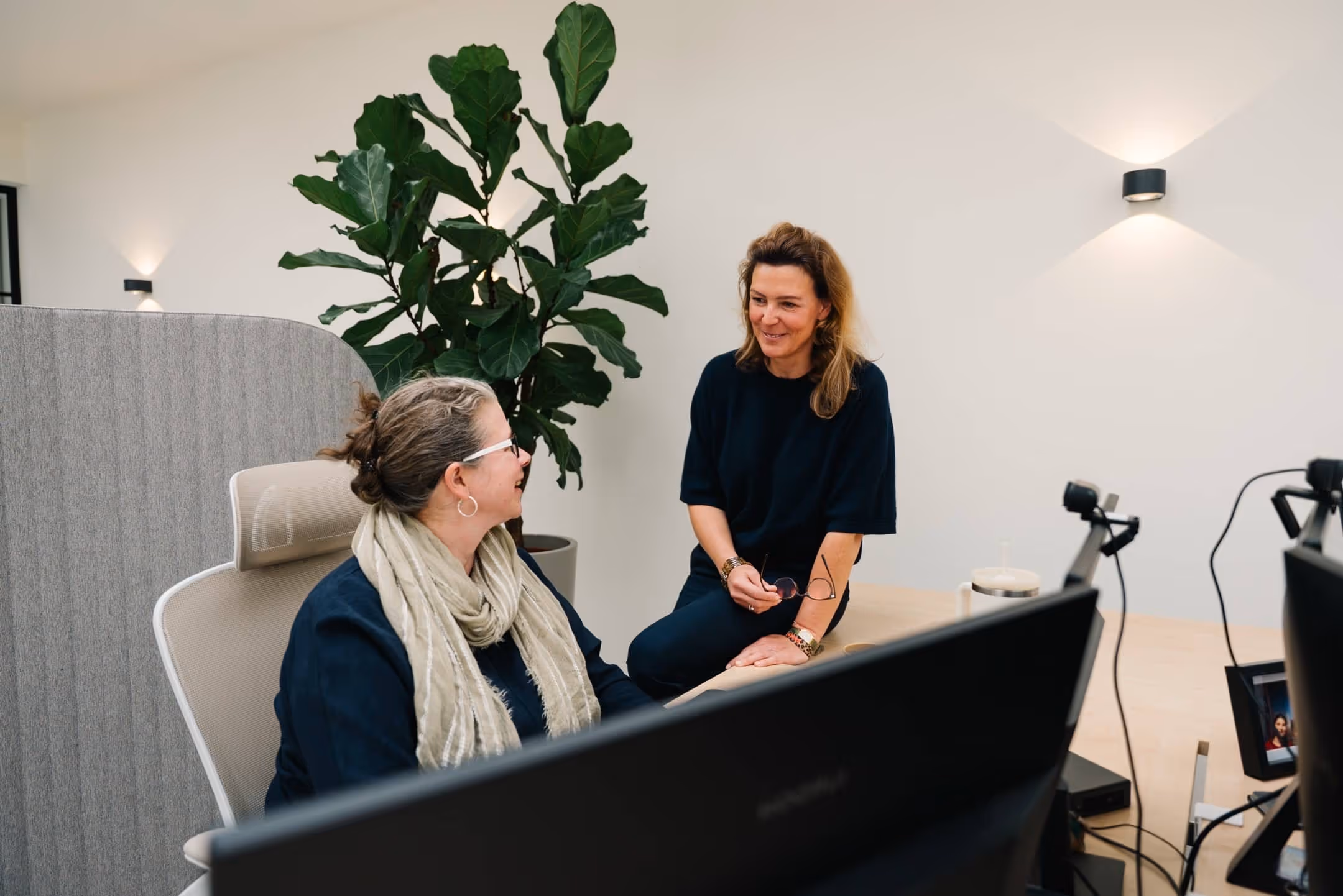 Zwei Frauen unterhalten sich freundlich in einem modernen Büro mit Pflanzen und Computern.
