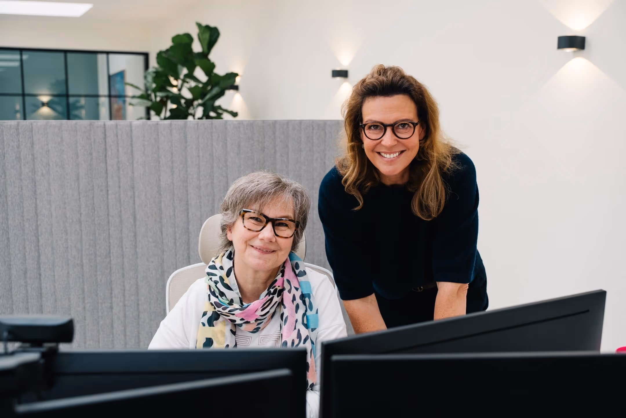 Zwei lächelnde Frauen mit Brille arbeiten gemeinsam an Computern in einem modernen Büro.