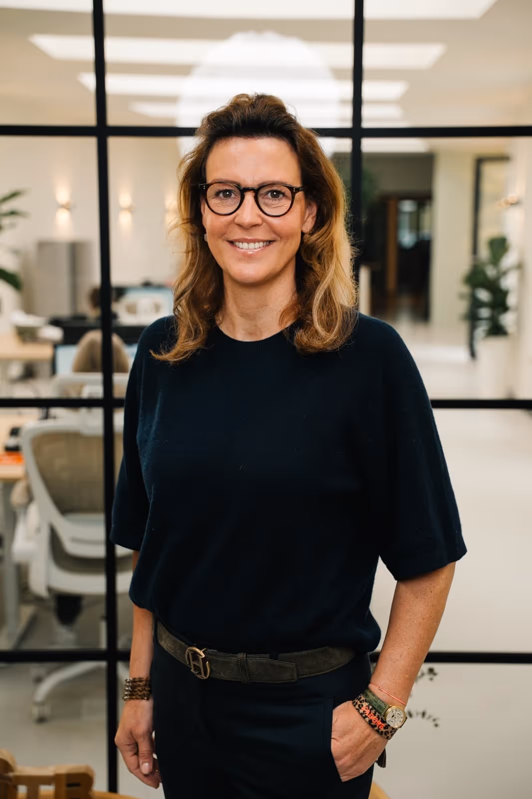 Frau mit Brille, dunkelblauem Oberteil und Armbändern steht vor einer Glaswand in einem modernen Büro.