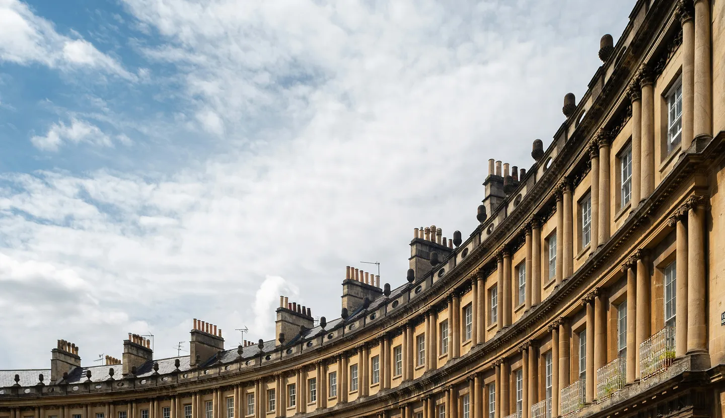 Curved Georgian townhouses built from honey-coloured Bath stone line a sweeping crescent beneath a blue sky filled with soft, streaked clouds.