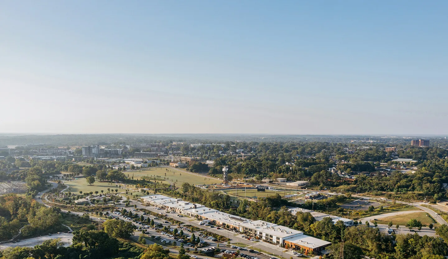 An aerial view of a suburban landscape with tree-lined streets, commercial buildings, sports fields, and parks stretching towards the horizon beneath a clear blue sky.