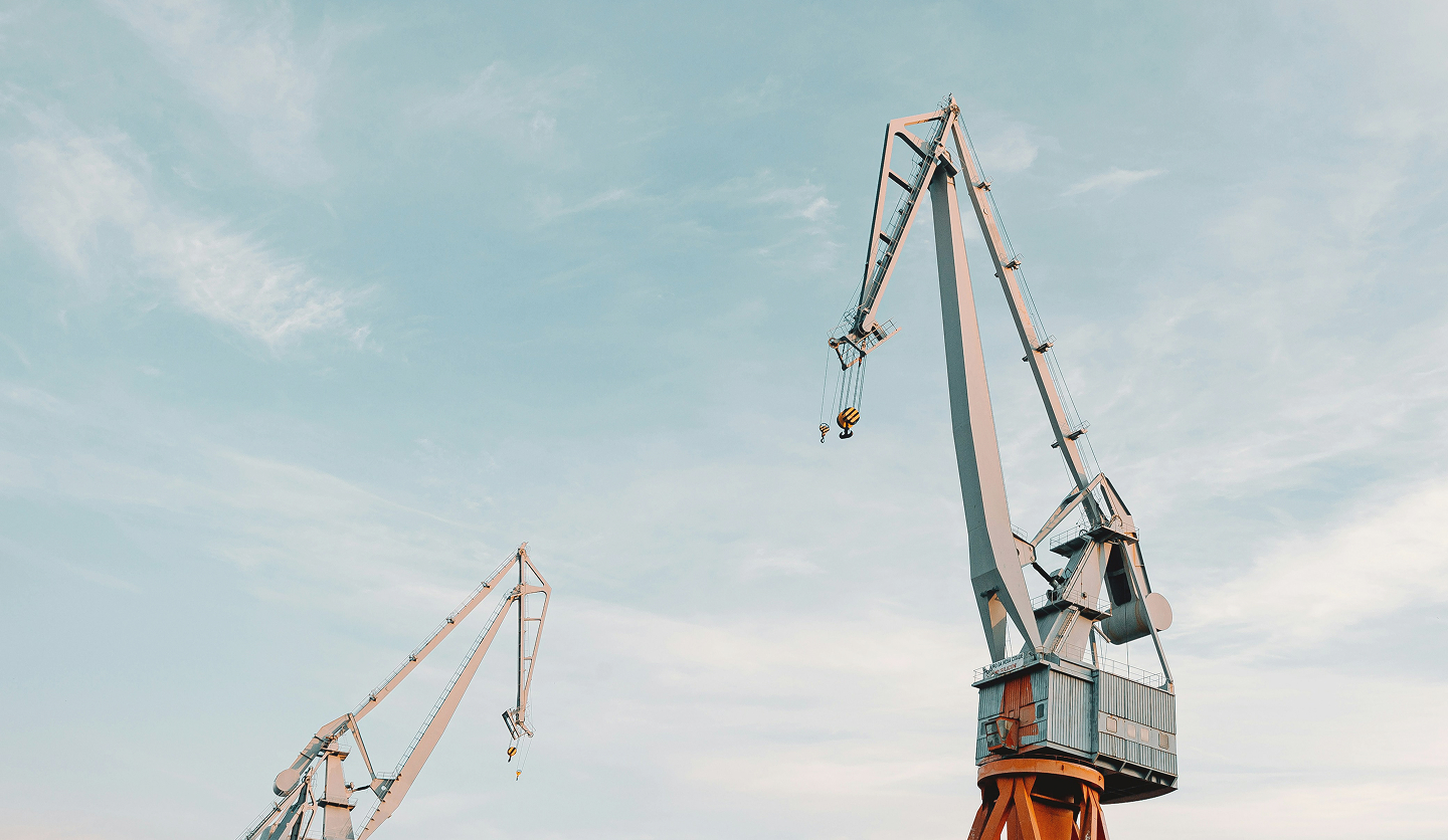 Two large industrial harbour cranes tower above the waterfront, silhouetted against a pale blue sky streaked with soft clouds.