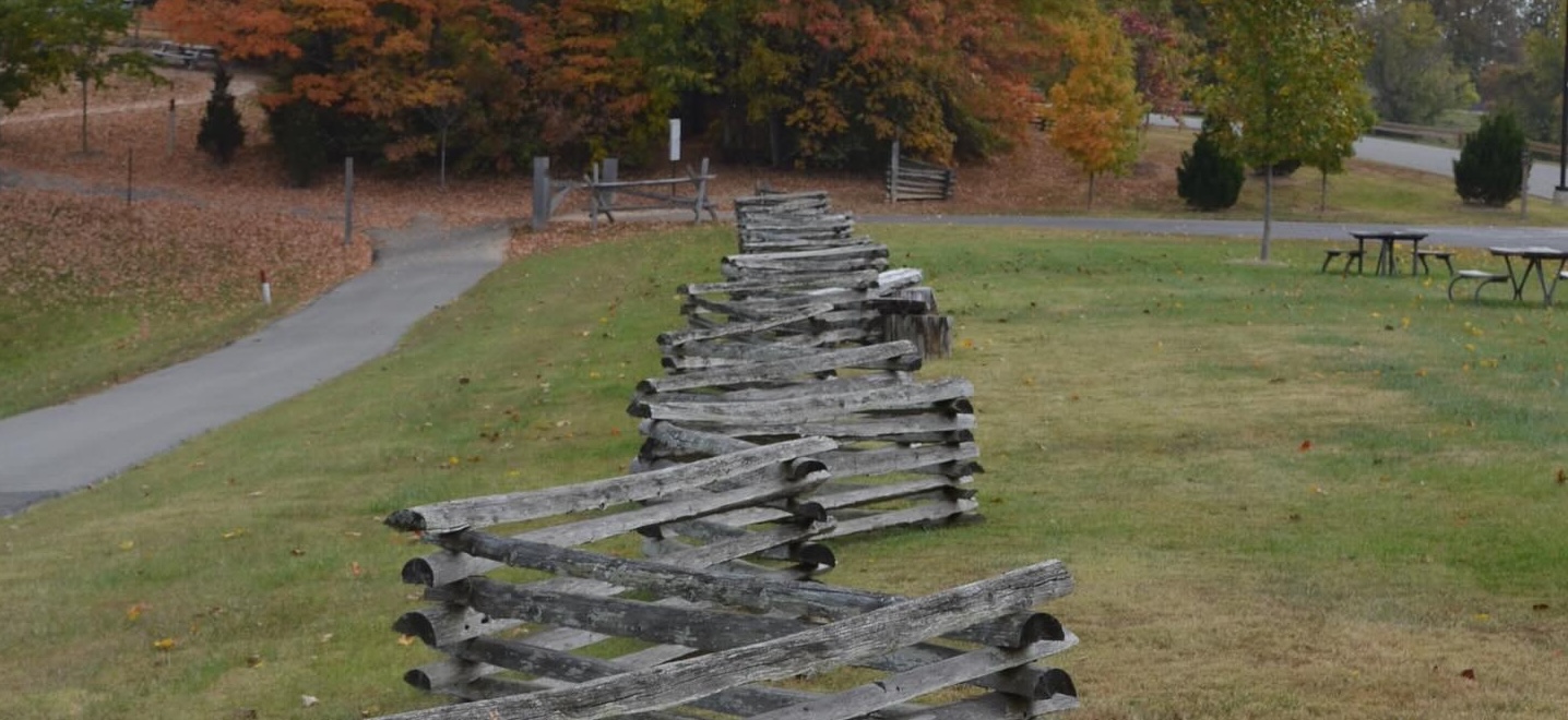 Rustic zigzag wooden fence on a grassy hill with autumn trees and picnic tables in the background.