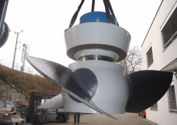 Large industrial propeller suspended by straps outdoors near buildings and vehicles.
