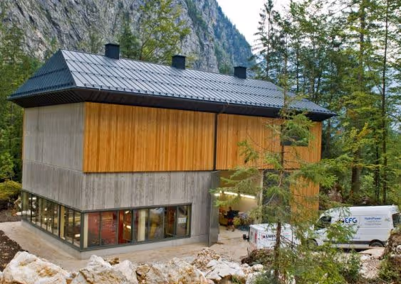 Modern two-story building with concrete and wood panels surrounded by trees and mountains, with two white vans parked nearby.
