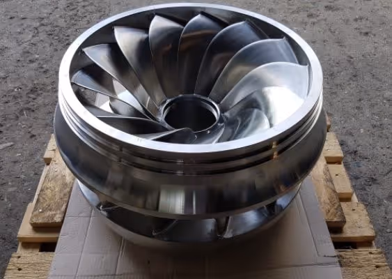 Polished metal turbine impeller with curved blades resting on a wooden pallet.