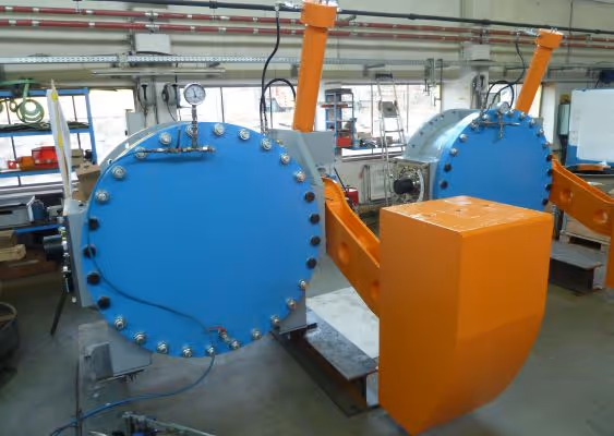 Industrial machinery with two large blue cylindrical chambers and orange mechanical arms inside a workshop.