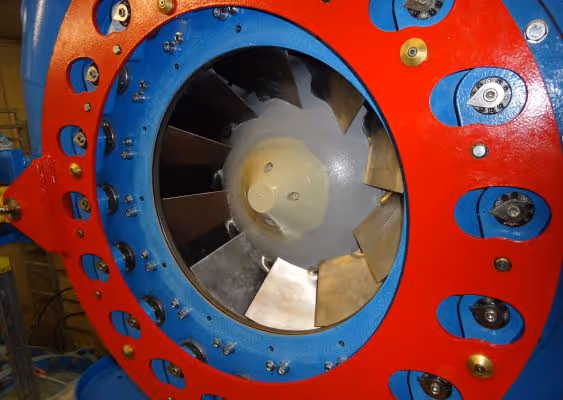 Close-up of a mechanical component featuring a circular metal fan or turbine enclosed by a red and blue housing with bolts and fittings.