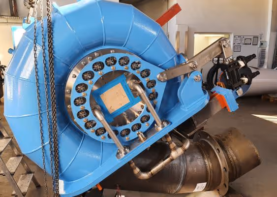 Close-up of a large blue industrial turbine or pump in a workshop, showing metal pipes, bolts, and mechanical components.