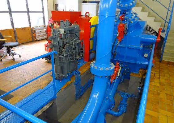 Industrial blue machinery with red valves and a gray control panel inside a facility with tiled floor and metal railings.