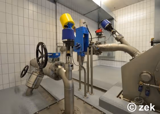 Industrial room with stainless steel pipes, valves, and control devices mounted on a tiled wall.
