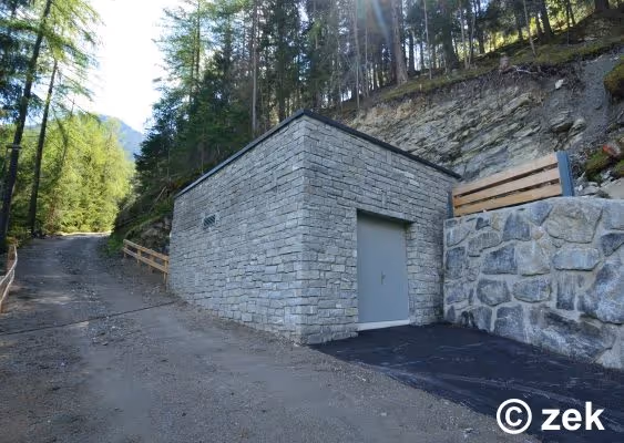 Small stone utility building with a closed gray door beside a gravel path surrounded by trees and rocky hillside.