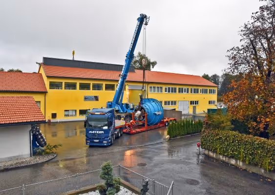 Blue crane lifting a large blue cylindrical tank onto a red flatbed trailer in front of a yellow industrial building on a wet paved area.