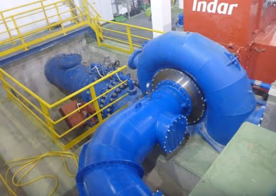Large blue industrial pump system with yellow safety railings in a facility with machinery and a red panel labeled 'Indar'.