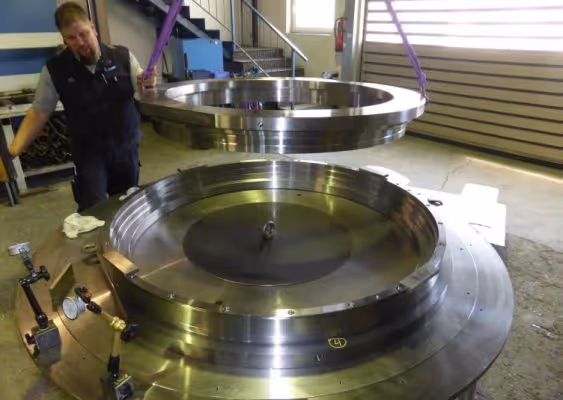 Worker assembling a large precision machined stainless steel ring using lifting straps in an industrial workshop.