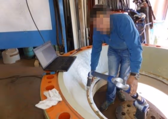 Person wearing blue shirt and jeans inspecting a large circular industrial component using a handheld device, next to an open laptop.