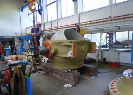 Industrial workshop with a large green machinery component suspended by an orange crane, with two workers nearby and windows in the background.
