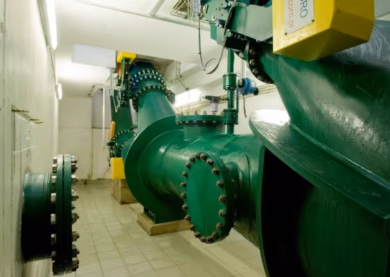 Large green industrial pipe system with flanges and valves inside a tiled room.