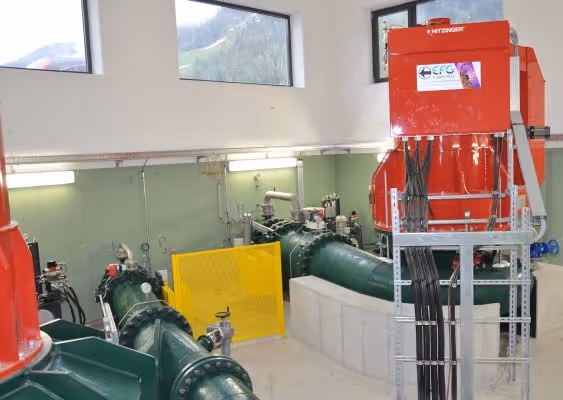 Interior of a hydropower facility with large green pipes and red control units under natural light from windows.