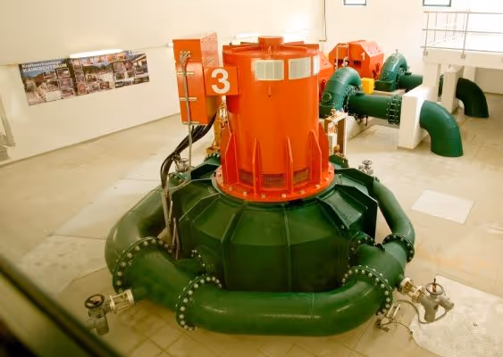 Interior view of a hydropower plant showing green and red turbine machinery with pipes and control valves.