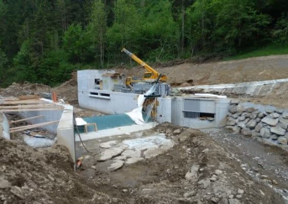 Partially constructed dam structure with concrete walls, heavy machinery, and surrounding earth in a forested area.