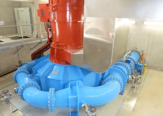 Industrial hydroelectric turbine with large blue pipes and a red cylindrical section in a clean mechanical room.