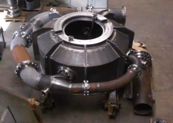 Large industrial metal machine with circular central opening and connected curved pipes on a workshop floor.
