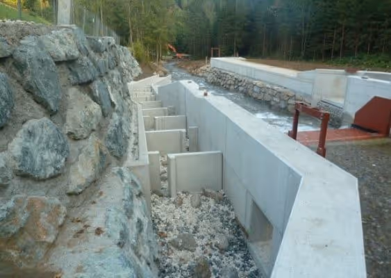 Concrete flood control barriers installed alongside a rocky embankment in a forested area.