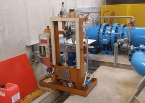 Industrial machinery setup with pipes, gauges, and a blue cylindrical component in a concrete room.