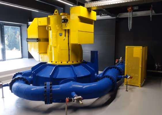 Large industrial machine with prominent blue curved pipes and a yellow mechanical unit inside a facility with dark walls and a window.