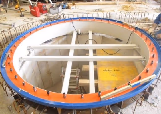 Large circular metal frame with bolts on a construction site, surrounding an open pit with scaffolding and equipment inside.