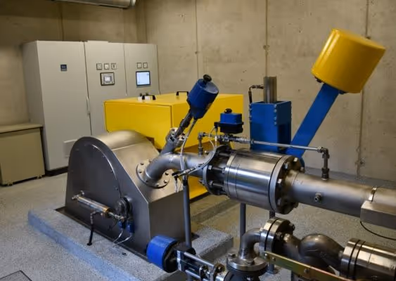 Industrial machinery with stainless steel components, blue and yellow instruments, and a control panel in a concrete room.